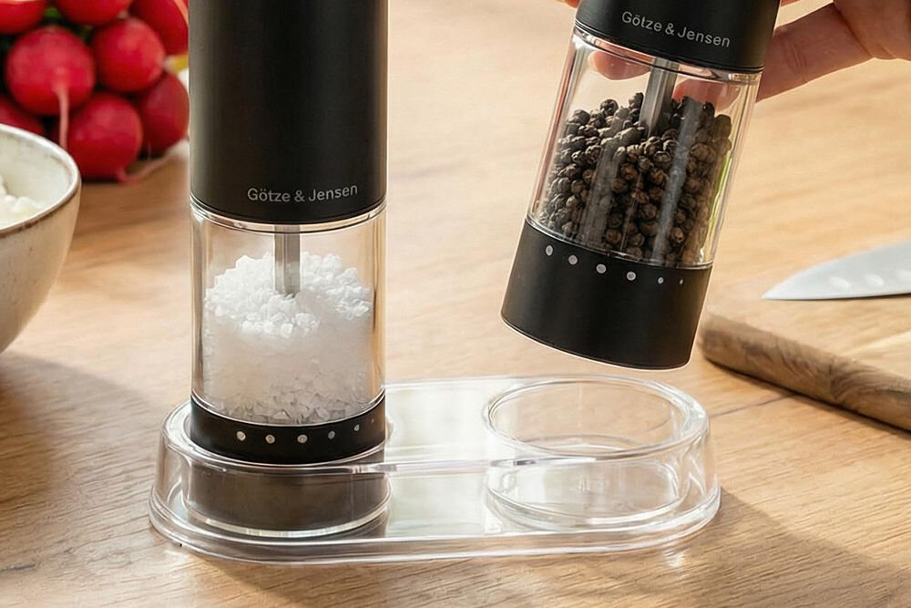 Zestaw młynków do przypraw GÖTZE & JENSEN KA420 Przezroczysta tacka ułatwiająca zachowanie czystości na blacie, z której wyjmowany jest czarny młynek elektryczny Götze & Jensen wypełniony ziarnami pieprzu.