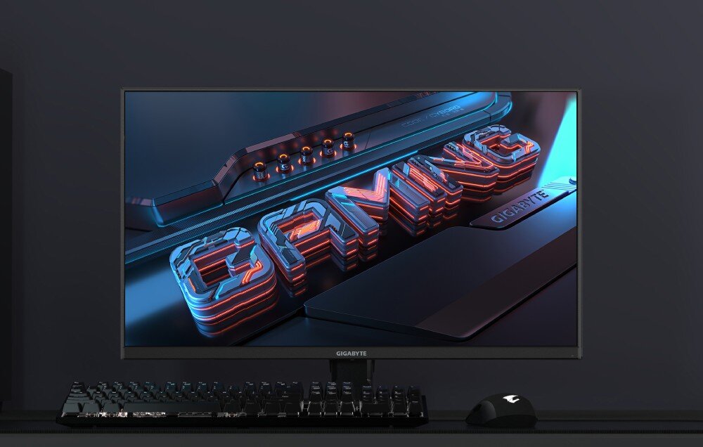 Monitor GIGABYTE M27UA Urządzenie zaprezentowane na stanowisku gracza wraz z podświetlaną klawiaturą i myszą, wyświetlające na ekranie renderowaną grafikę z napisem 'GAMING' i mechanicznymi detalami, opis