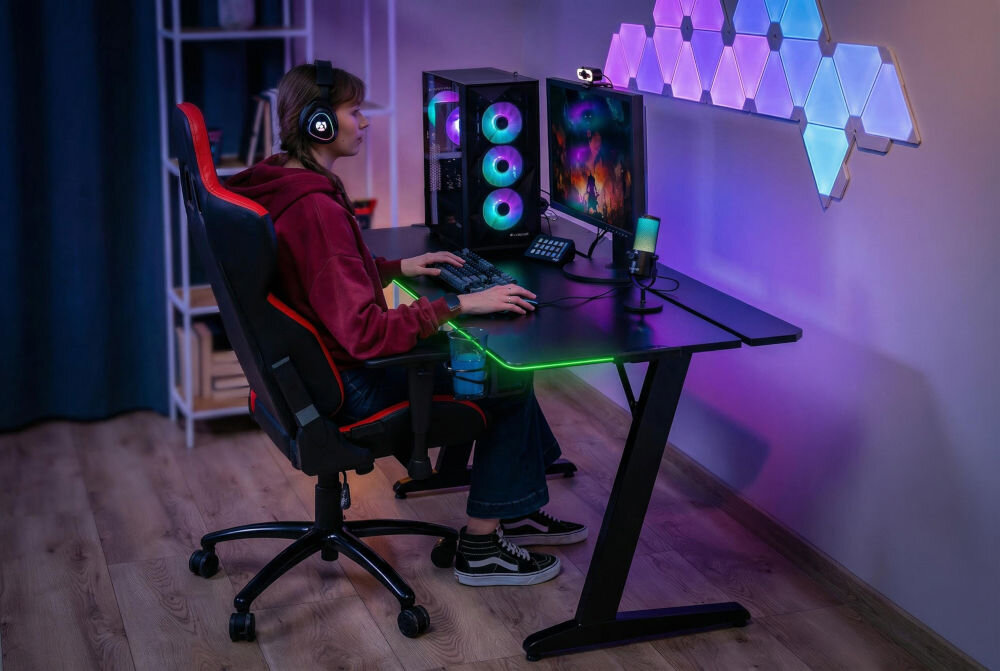 Biurko MAD DOG GD100K Kobieta siedząca przy czarnym biurku gamingowym MAD DOG z podświetleniem LED RGB, na którym stoi komputer stacjonarny, monitor i mikrofon.