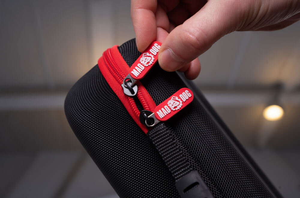 Etui MAD DOG PPA500K dla PlayStation Portal z szkłem hartowanym i ściereczką w zestawie Zbliżenie na dłoń trzymającą dwa czerwono-czarne uchwyty zamków błyskawicznych, na których widnieje logo MAD DOG. Zamki są wpięte w czerwony zamek czarnego, twardego etui na konsolę.