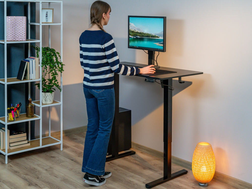 Biurko XQUANTUM ED100K Elektronicznie regulowane biurko XQUANTUM w pozycji stojącej przy białej ścianie z regałem na książki