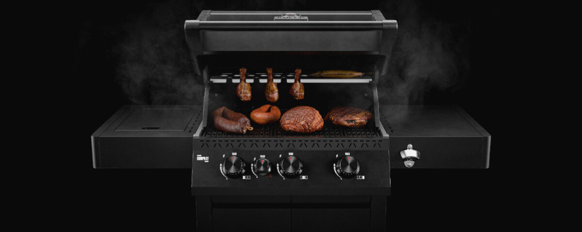 Czarny grill gazowy z otwartą pokrywą, na ruszcie znajdują się różne rodzaje mięsa. Na panelu przednim widoczne są pokrętła do regulacji temperatury.