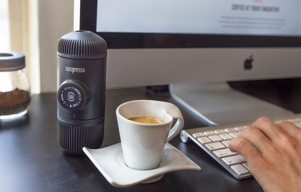 Na biurku stoi szary ekspres Nanopresso obok filiżanki z kawą na białym spodku. W tle widać monitor i klawiaturę, a ręka dotyka klawiszy.