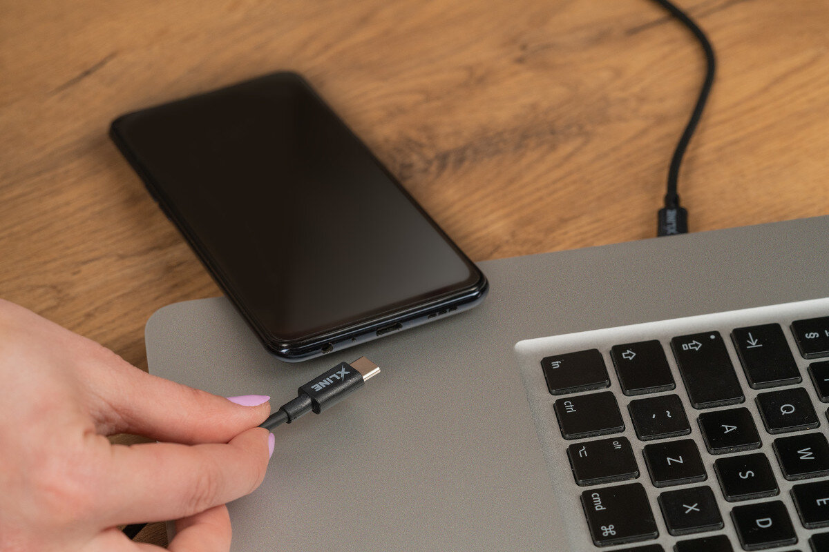 Osoba trzyma czarny kabel USB-C z logo \'XLINE\', który jest podłączany do laptopa. Obok znajduje się smartfon na drewnianym blacie.