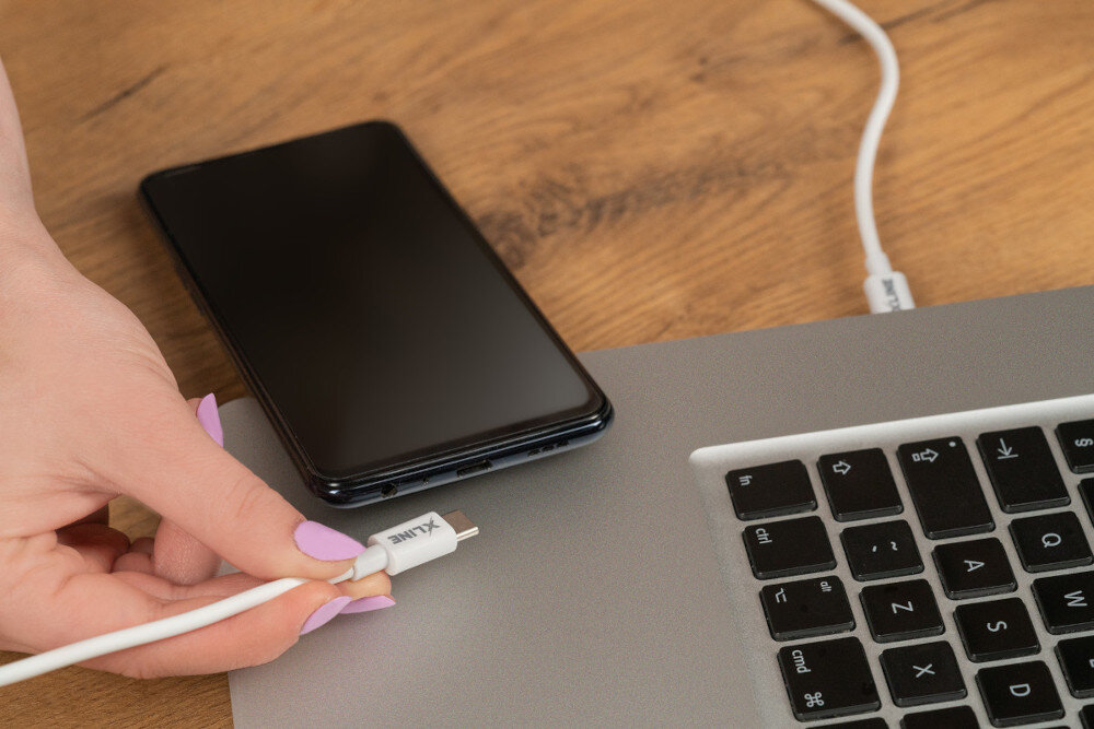 Osoba trzyma biały kabel USB-C z logo \'XLINE\', który jest podłączany do laptopa. Obok znajduje się czarny smartfon na drewnianym blacie.