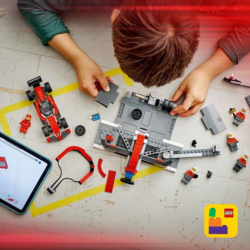 Dziecko składa zestaw klocków LEGO, w tym czerwony bolid i elementy pit stopu. Na stole widoczne są także figurki mechaników oraz tablet z instrukcją budowy.