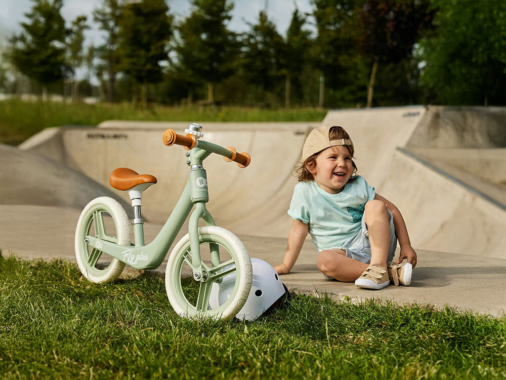 Rowerek biegowy KINDERKRAFT Fly Plus Różowy dziecko siedzi na trawie obok stoi miętowy rowerek jasne koła gumowe uchwyty dzwonek brązowe siedzisko świetna zabawa aktywne wspieranie rozwoju dziecka dziecko doskonali koordynację ruchową wzmacnia mięśnie nóg uczy się utrzymywać równowagę budowanie zdrowego nawyku codziennej aktywności przyjemność wartościowy trening 