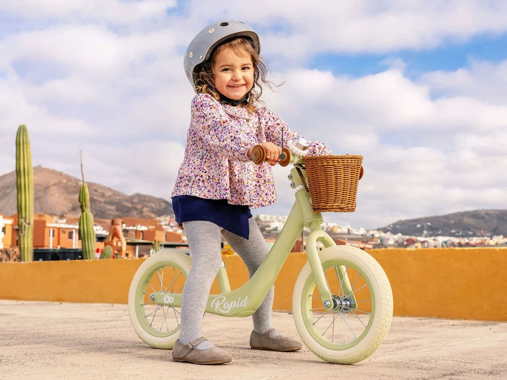 Rowerek biegowy KINDERKRAFT Rapid 2 Różowy uśmiechnięta dziewczynka w kasku jedzie na zielonym rowerku gumowe uchwyty brązowy koszyk jasne koła dla dzieci od 2 roku życia wygoda bezpieczeństwo umiejętności motoryczne solidna rama stabilność maksymalne obciążenie do 35 kg dostosowuje się do rosnących potrzeb pierwsze rowerowe przygody wspiera rozwój koordynacji i równowagi  