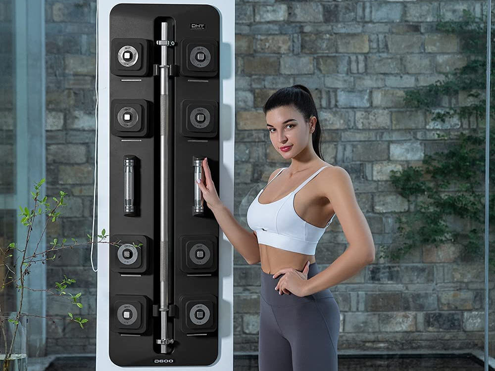 Zestaw hantli S-LINE SA032 2 x 10 kg + sztanga 121 cm pomieszczenie osoba stoi przy ścianie na której zawieszony jest zestaw w tle ściana z szarej cegły trening siłowy w domu regulowane obciążenie