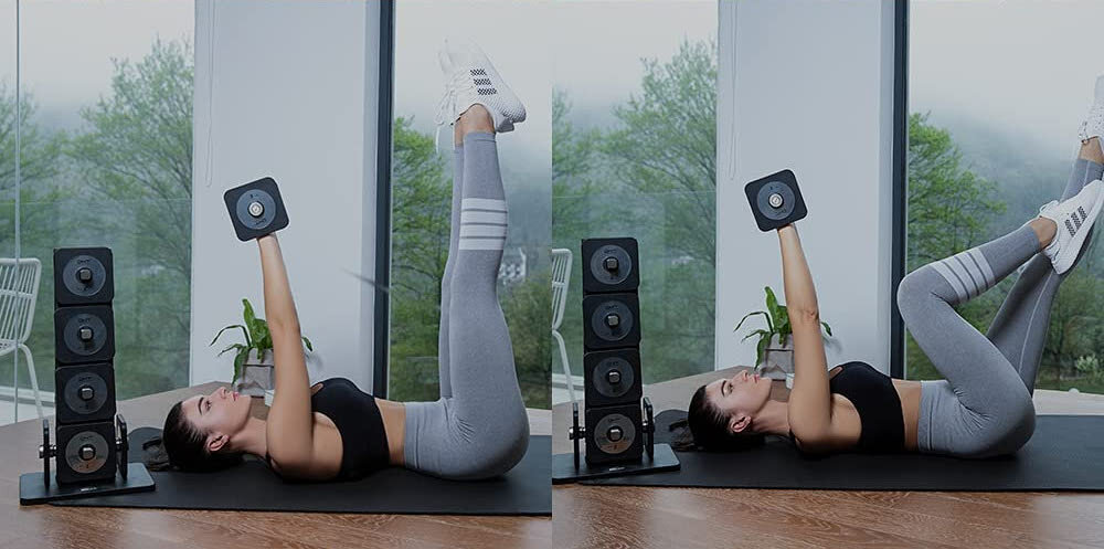 Osoba ćwiczy na macie z hantlami w rękach, w tle widoczne są duże okna z widokiem na drzewa. Obok znajduje się stojak z dodatkowymi hantlami.