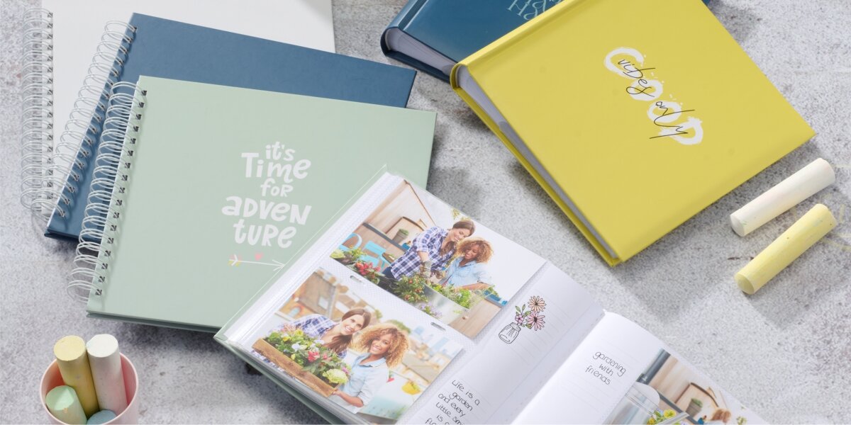 Na obrazie widoczne są różne albumy fotograficzne, w tym zielony z napisem \'it\'s Time for adventure\'. Obok albumów leżą kolorowe kredy.