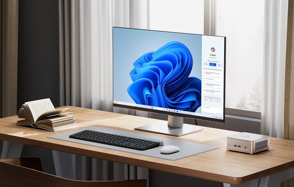 Komputer MINISFORUM UM870 Plus Srebrny mini PC na drewnianym biurku obok monitora z systemem Windows i otwartej książki.