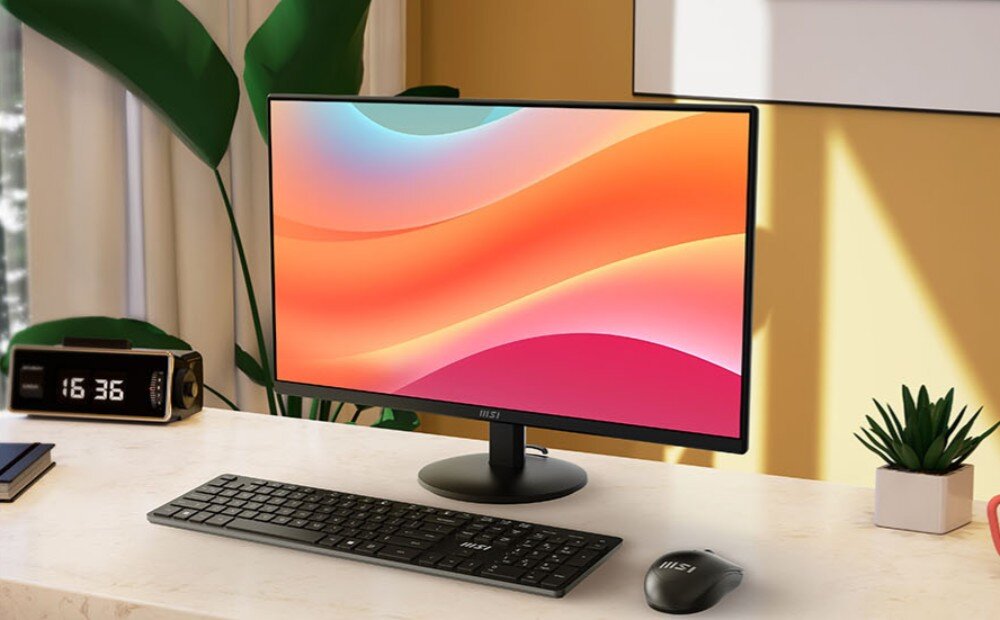 Monitor MSI Pro MP272L Monitor na biurku w domowym wnętrzu, z klawiaturą i myszką przed nim oraz zegarem cyfrowym pokazującym godzinę 16:36, opis
