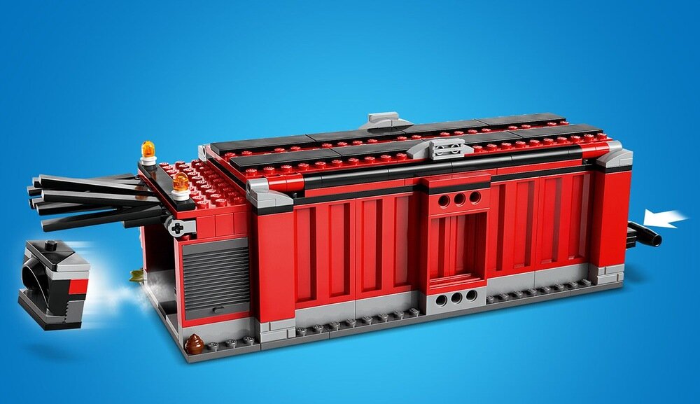 Na niebieskim tle znajduje się czerwony model kontenera z klocków, z czarnymi i szarymi elementami oraz widocznymi detalami przypominającymi światła ostrzegawcze.