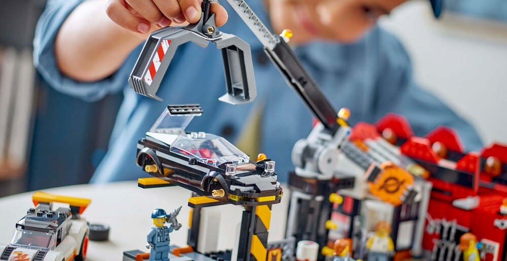 Na obrazie widać zestaw LEGO przedstawiający złomowisko z samochodami, z dźwigiem podnoszącym czarny samochód. W tle znajduje się dziecko bawiące się zestawem.