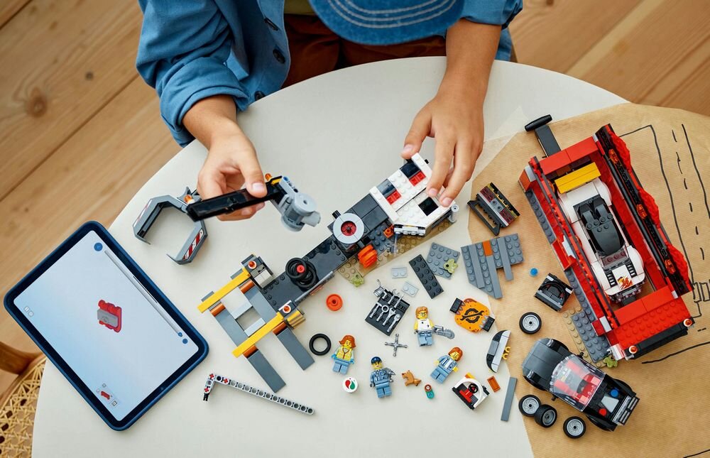Na stole znajdują się elementy zestawu LEGO, w tym części pojazdów i minifigurki. Obok leży tablet z wyświetlonym obrazem klocka LEGO.