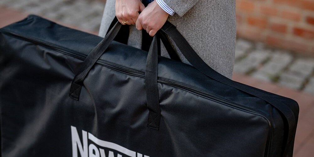 Zbliżenie na dłonie kobiety w szarym płaszczu trzymającą torbę transportową z napisem Newell, na tle ceglastego budynku.