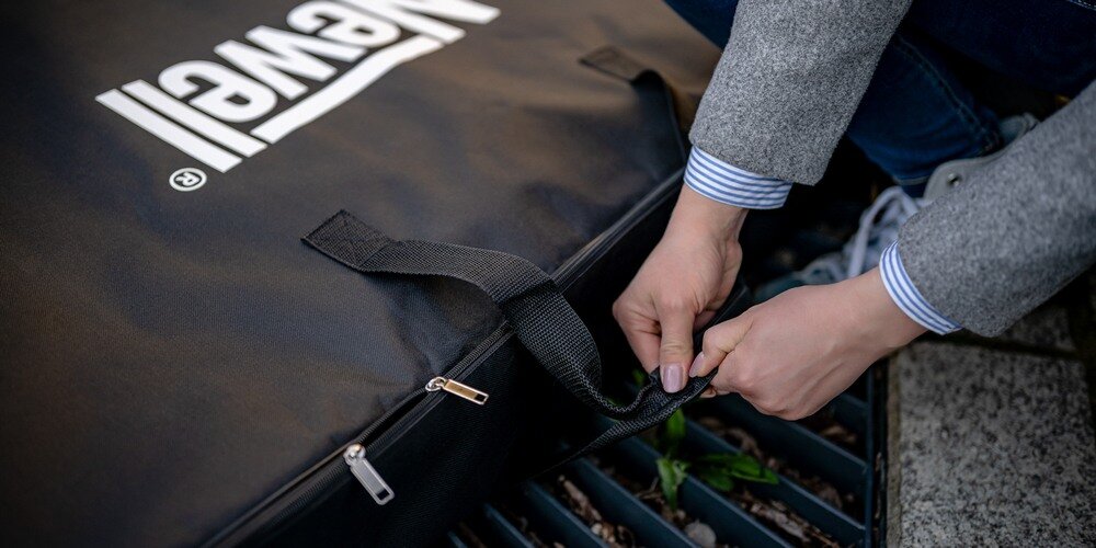 Zbliżenie na dłonie kobiety w szarym płaszczu łączącą uchwyty przed podniesieniem torby Newell leżacej na kratce na ulicy.