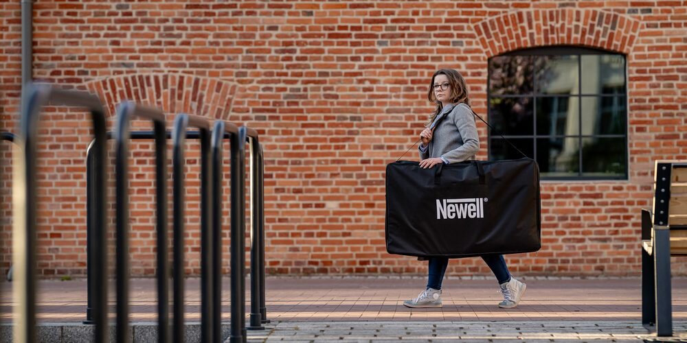 Kobieta w szarym płaszczu na tle ceglanego budynku niesie na ramieniu dużą czarną torbę transportową Newell.