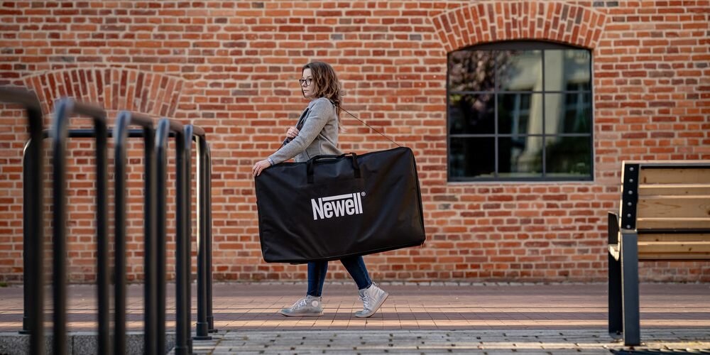 Kobieta idzie wzdłuż ceglastego budynku, niosąc na ramieniu torbę Newell. Po lewej metalowe barierki, po prawej ławeczka.