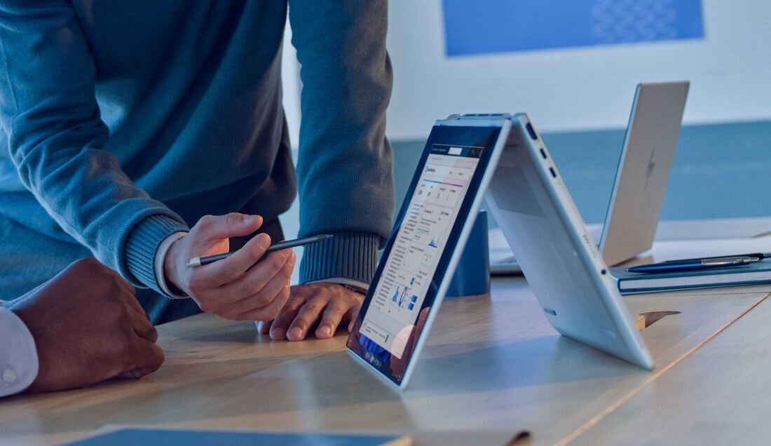 Laptop HP EliteBook X Flip w trybie namiotu na drewnianym biurku w nowoczesnym wnętrzu, ekran z aplikacją do analizy danych i wykresami, w tle drugi laptop i akcesoria biurowe w niebieskim oświetleniu. - Wi-Fi 7 Bluetooth 5.4
