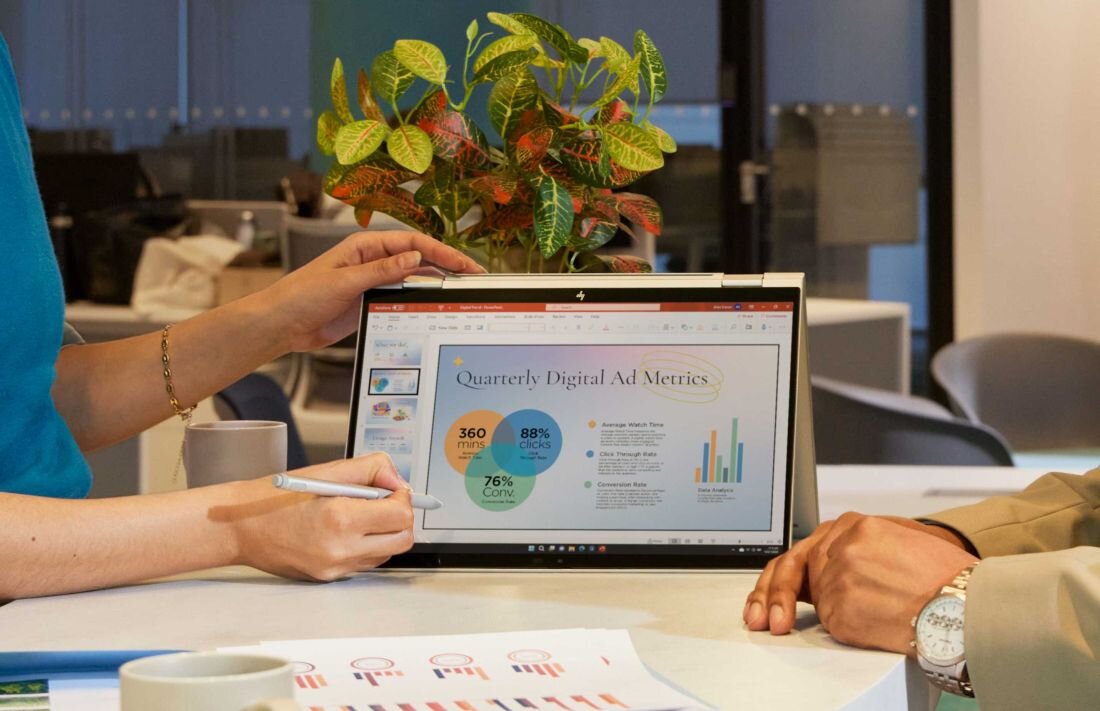 Kobieta w niebieskim swetrze trzymająca piórko cyfrowe podczas pracy na laptopie HP EliteBook X Flip w trybie tabletu, na ekranie prezentacja z wykresami Quarterly Digital Ad Metrics, na białym biurku dokumenty z kolorowymi wykresami i kubek kawy. - Bateria o pojemności 68 Wh, HP Power Manager