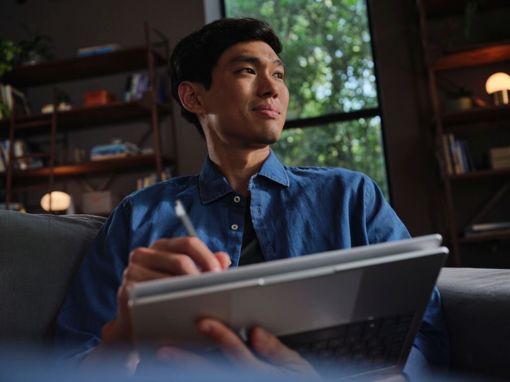 Młody mężczyzna w niebieskiej koszuli uśmiechający się podczas pracy na laptopie HP EliteBook X Flip w trybie tabletu z piórkiem cyfrowym, w przytulnym wnętrzu z półkami książek i ciepłym oświetleniem. - Srebrna elegancka obudowa, wymiary   