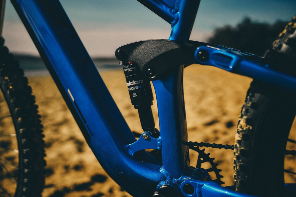 Zbliżenie na niebieską ramę roweru z widocznym amortyzatorem marki RockShox. W tle widać piaszczystą plażę.