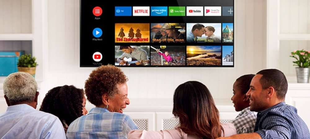 Na obrazie widać telewizor z wyświetlonym interfejsem Android TV, na którym znajdują się ikony aplikacji takich jak Netflix, YouTube i Prime Video. Przed telewizorem siedzi grupa osób, patrzących na ekran.