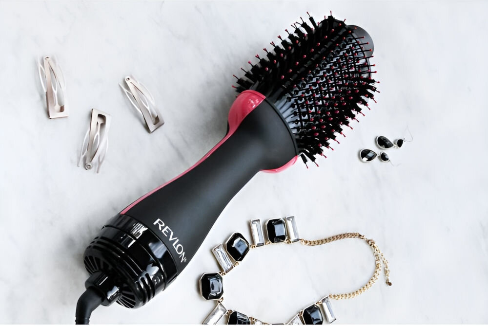 Na obrazie widoczna jest suszarko-lokówka z napisem \'REVLON\' na czarnej rączce, otoczona biżuterią i spinkami do włosów. Produkt ma różowe akcenty i okrągłą szczotkę z czarnymi i czerwonymi włoskami.
