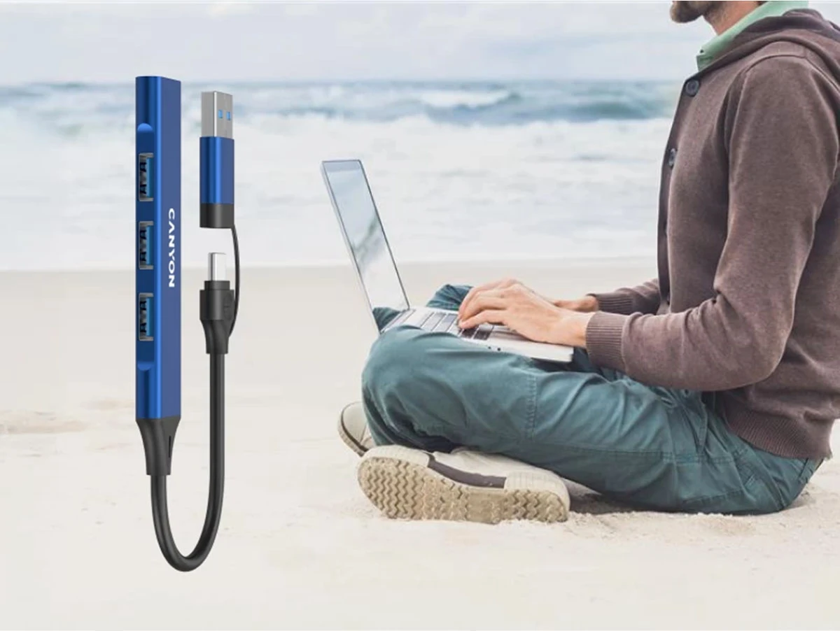 Na obrazie widoczny jest niebieski hub USB z trzema portami i logo \'CANYON\'. W tle osoba siedzi na plaży z laptopem na kolanach.