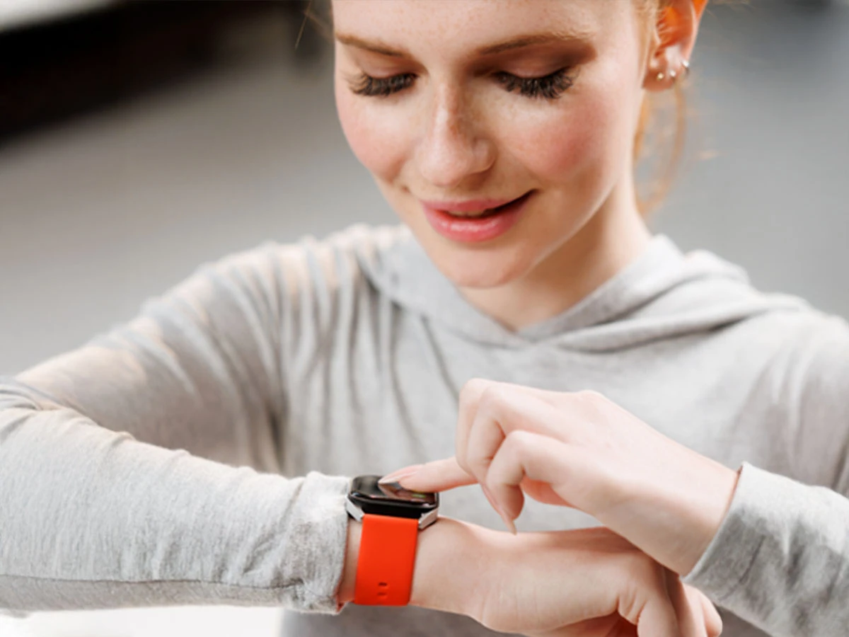 Osoba w szarej bluzie dotyka ekranu smartwatcha z czerwonym paskiem na nadgarstku. Smartwatch ma srebrną obudowę.