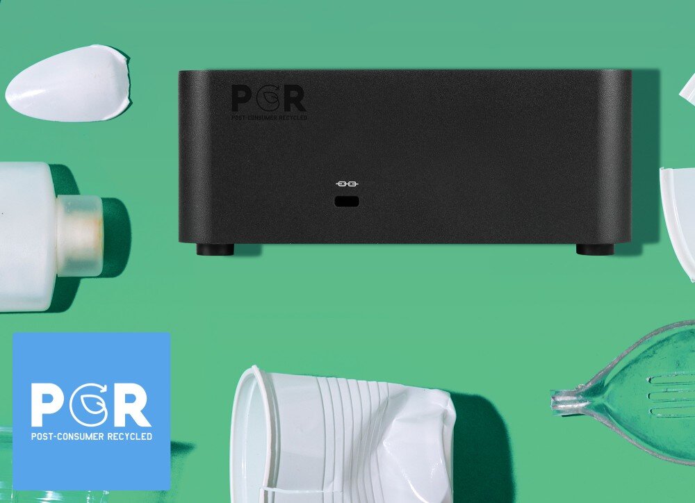 Komputer MSI Cubi NUC AI+ 2MG-007EU Ciemnoszary komputer z napisem 'PCR Post-Consumer Recycled' ustawiony na tle zielonej powierzchni z rozrzuconymi plastikowymi odpadami i ikoną recyklingu, materiały, recykling