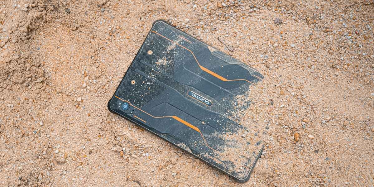Tablet leży na piasku, częściowo pokryty ziarnami. Na obudowie widoczne są pomarańczowe akcenty i logo \'OUKITEL\'.