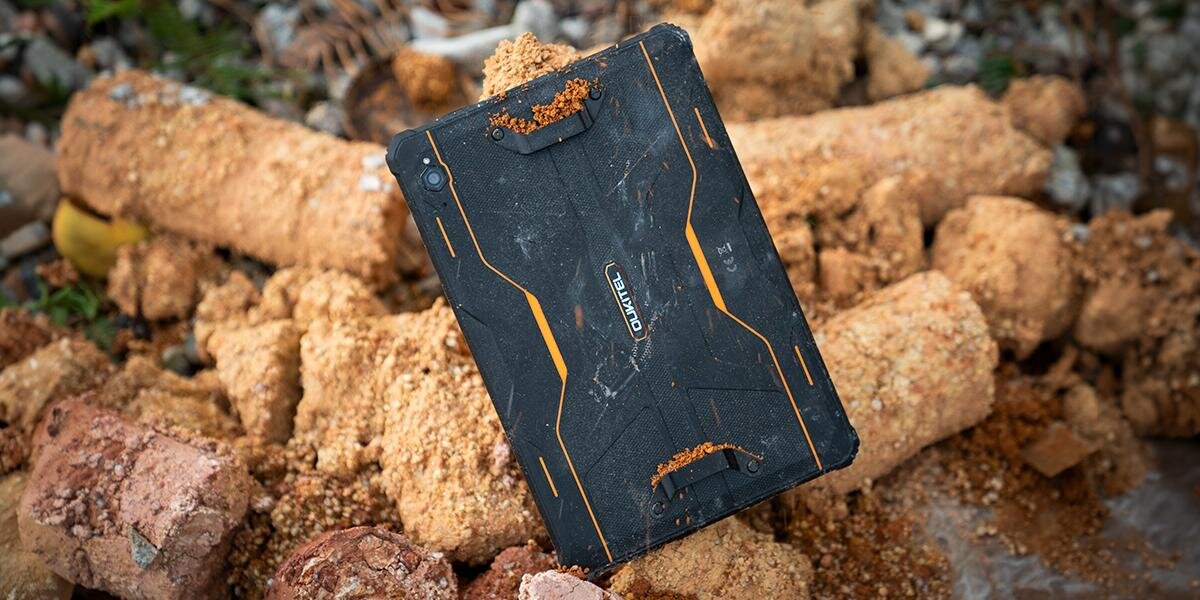Tablet w czarnej obudowie z pomarańczowymi akcentami leży na ziemi pokrytej piaskiem i kamieniami. Na obudowie widoczne jest logo \'OUKITEL\'.