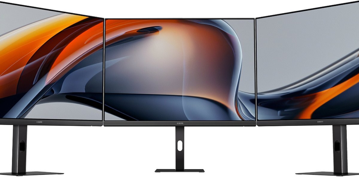 Trzy monitory z cienkimi ramkami, wyświetlające abstrakcyjną grafikę w odcieniach pomarańczowego i szarego. Na dolnej ramce każdego monitora widoczne jest logo \'XIAOMI\'.