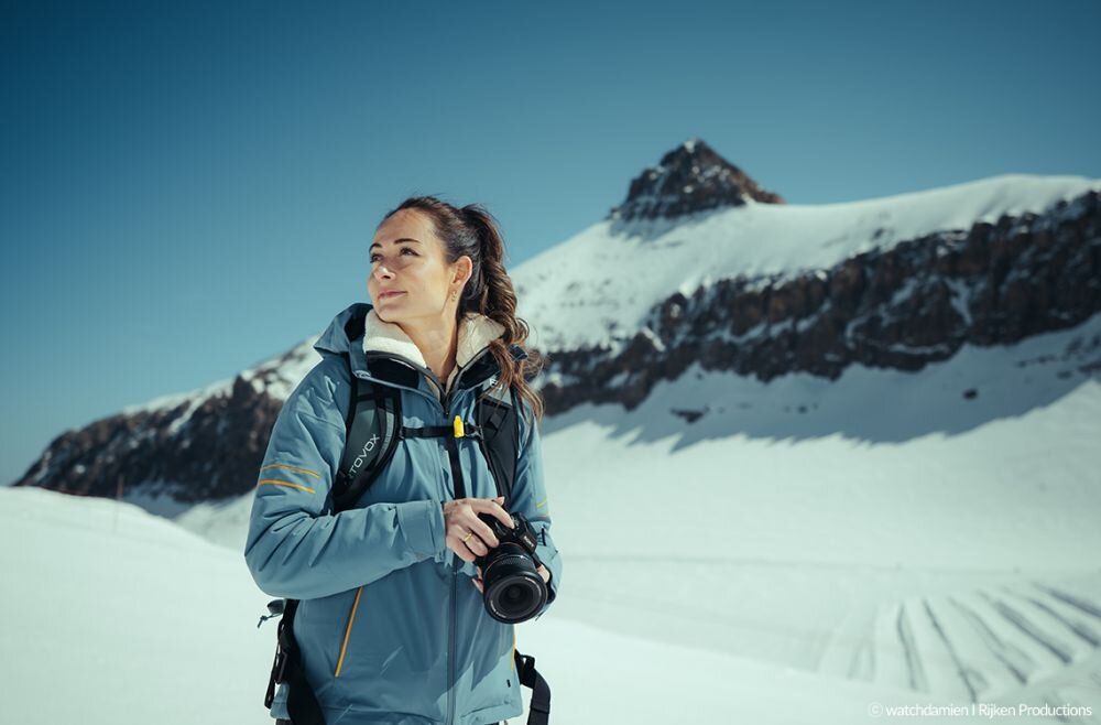 Osoba w niebieskiej kurtce trzyma aparat fotograficzny na tle ośnieżonych gór. W prawym dolnym rogu widoczny jest napis: © watchwomen | Rijken Productions.