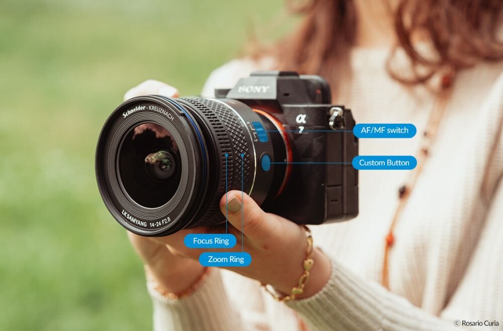 Osoba trzyma aparat Sony z zamontowanym obiektywem Samyang AF 14-24 mm f/2.8. Na obiektywie widoczne są oznaczenia: \'Focus Ring\', \'Zoom Ring\', \'AF/MF switch\', \'Custom Button\'.