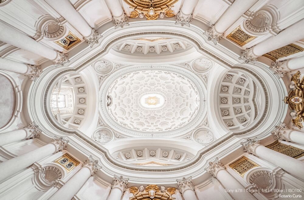 Widok na bogato zdobiony sufit z białymi i złotymi elementami architektonicznymi oraz kolumnami. Na dole obrazu widoczny jest tekst: \'Sony α7III | 14mm | f/11 | 1/80sec | ISO100 © Rosario Curia\'.