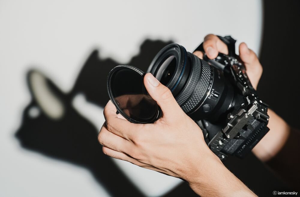 Osoba trzyma aparat fotograficzny z zamontowanym obiektywem, a w drugiej ręce trzyma filtr fotograficzny. Na ścianie widać cień aparatu i dłoni.