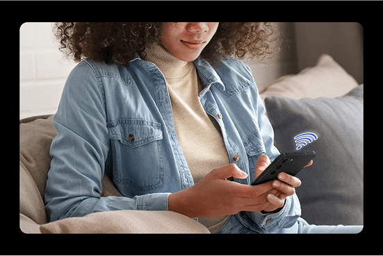 polacz z aplikacja Osoba siedzi na kanapie, trzymając w rękach smartfon z widoczną ikoną sygnału Wi-Fi. Ubrana jest w jasną koszulę jeansową i jasny sweter.