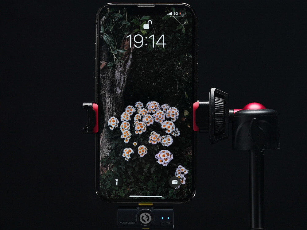System bezprzewodowy HOLLYLAND Lark M2 Combo Grafitowy prezentacja odbiornika podłączonego do telefonu stojącego na statywie czarne tło zdalne sterowanie rozpoczęcie i zatrzymanie nagrania wykonywanie zdjęć sprawna obsługa