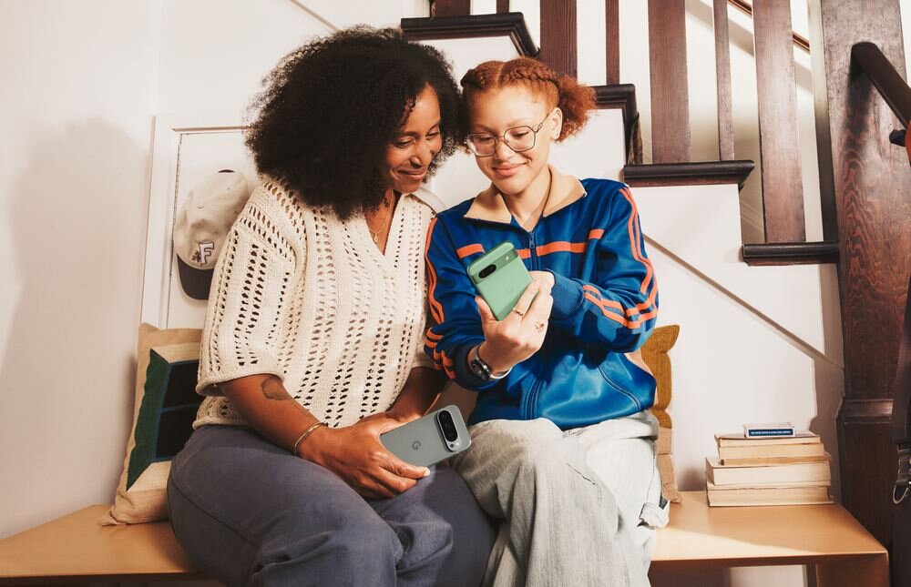 Dwie osoby siedzą na schodach, trzymając w rękach smartfony w różnych kolorach. W tle widać drewniane schody i kilka książek na półce.