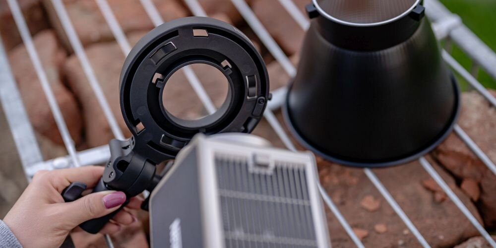 Dłoń trzyma czarny uchwyt montażowy Newell obok srebrnej lampy LED i czarnej czaszy na metalowej kratce.