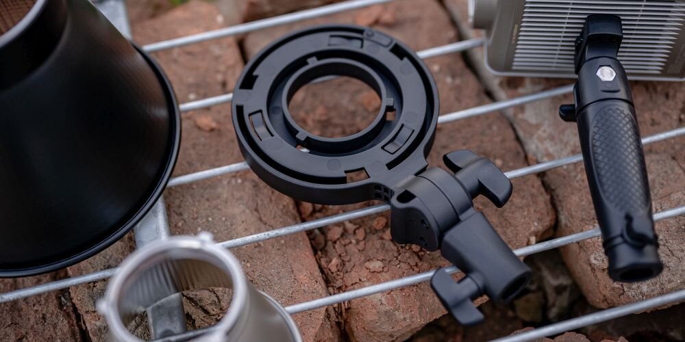 Czarny uchwyt montażowy z pokrętłami leży na metalowej kratce obok elementów lampy i ceglanego podłoża.