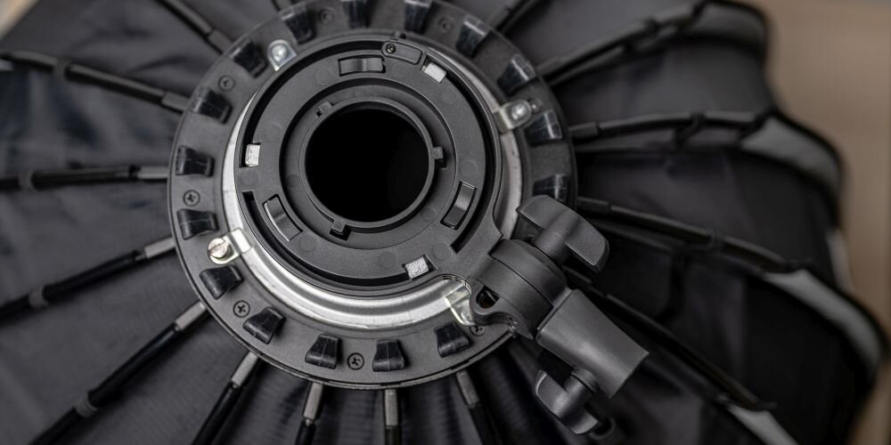 Centralny widok na czarny pierścień mocujący softbox z rozłożonymi prętami i pokrętłem regulacyjnym.