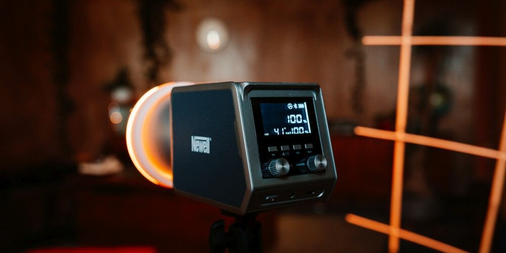 Lampa studyjna Newell na statywie w ciepłym, drewnianym wnętrzu. Duży okrągły pierścień LED świeci pomarańczowym światłem wokół kwadratowego panelu. Wyświetlacz pokazuje 100% mocy, temperaturę 41° i inne parametry. W tle drewniane belki i pomarańczowe linie świetlne.