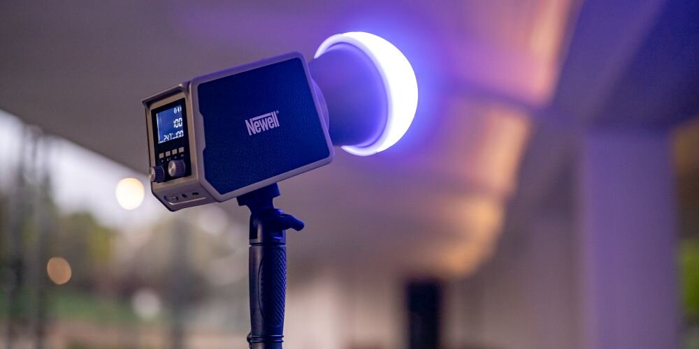 Lampa studyjna Newell na statywie w wieczornym świetle. Kwadratowy panel z niebieską obudową i dużym okrągłym pierścieniem LED świecącym fioletowo-niebieskim światłem. Wyświetlacz pokazuje 100% mocy, 24°, 100% i ikony. Tło to taras z balustradą i rozmyte światła.