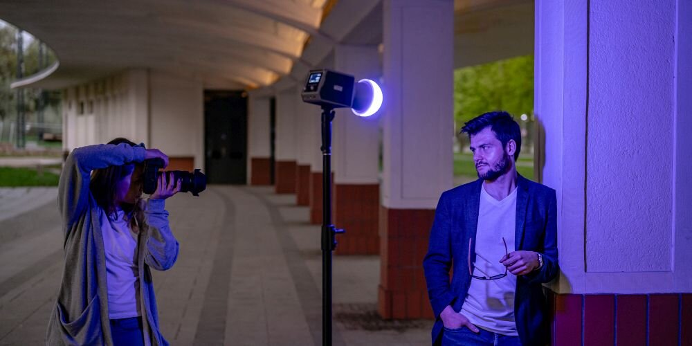 Kobieta w szarej bluzie i niebieskich jeansach fotografuje aparatem z dużym obiektywem, zasłaniając oczy dłonią. Obok na statywie lampa Newell świeci niebiesko-fioletowym światłem z okrągłym pierścieniem LED. Mężczyzna w granatowej marynarce opiera się o filar, trzymając okulary.