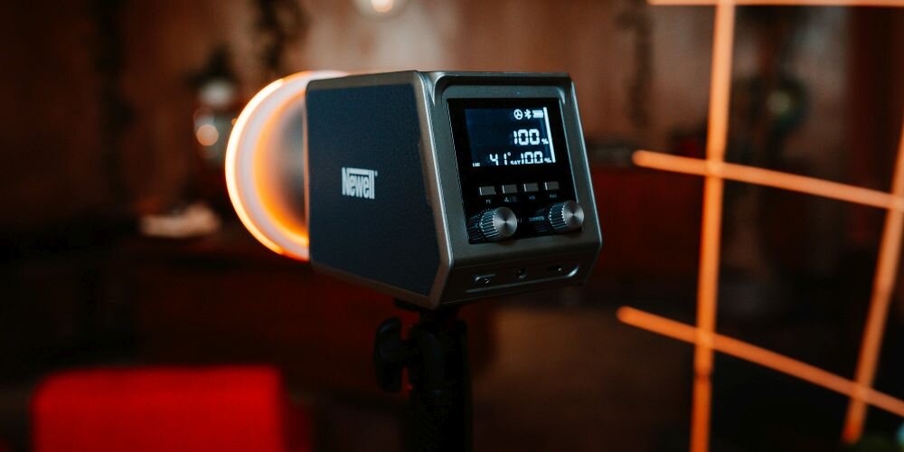Kompaktowa lampa LED na statywie, z pierścieniowym światłem, wyświetlaczem i pokrętłami sterowania, na ciemnym tle.